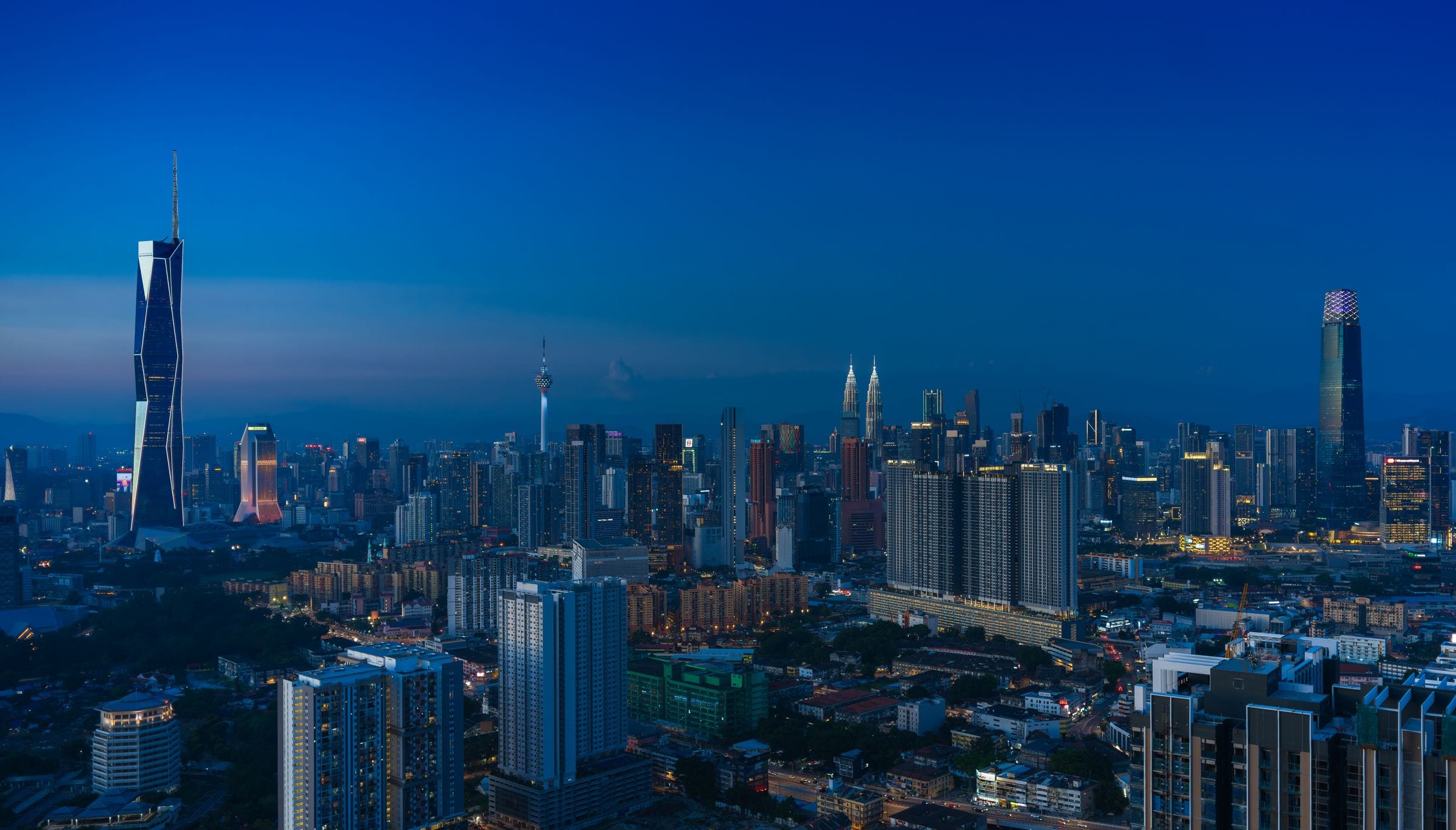 Panoramic blue-hour view of the Kuala Lumpur skyline with the Petronas Twin Towers and KL Tower in the distance among many buildings and evening city lights